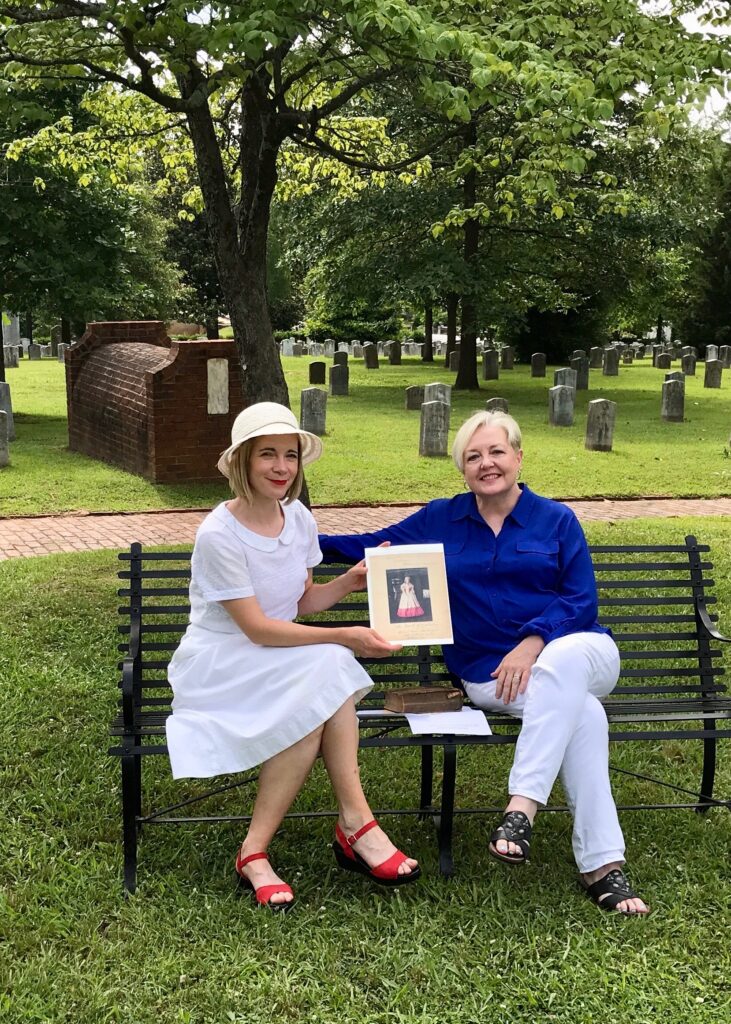 Karen Cox and Lucy Worsley on a bench in a cemetery.