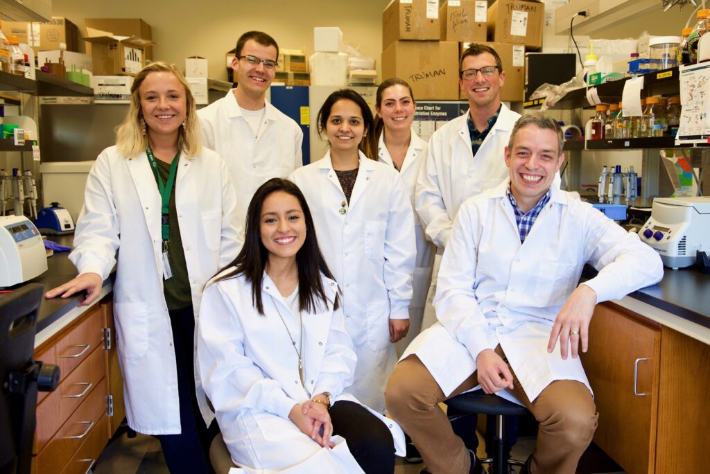 A group of 7 men and women in a lab in white lab coats.